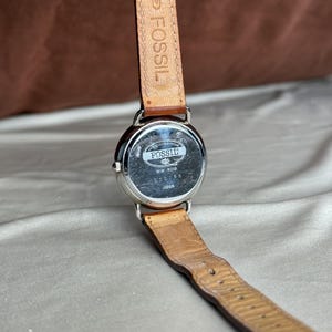 Puede incluir: Un reloj Fossil con correa de cuero marr&oacute;n. La esfera plateada muestra el logotipo de Fossil, el n&uacute;mero de modelo "WW-8318" y "Japan". La correa muestra desgaste.