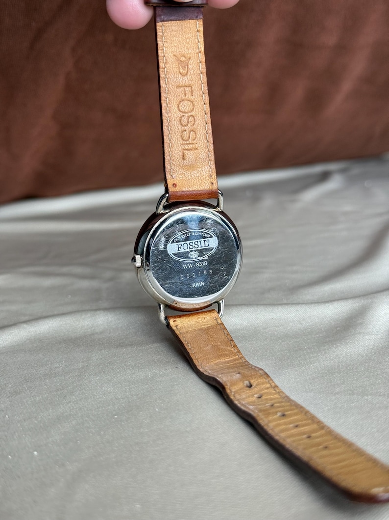 Puede incluir: Un reloj Fossil con correa de cuero marr&oacute;n. La esfera plateada muestra el logotipo de Fossil, el n&uacute;mero de modelo "WW-8318" y "Japan". La correa muestra desgaste.