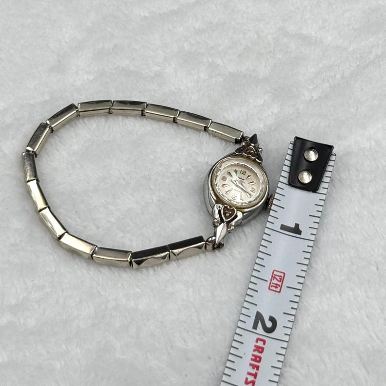 May include: A silver-toned vintage wristwatch with a rectangular link band. The watch face is round with a white background and black numerals. A measuring tape is next to the watch. The watch band is approximately 6 inches long.