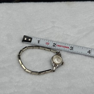 May include: A vintage silver-toned wristwatch with a small, round face and a linked band. The watch is next to a measuring tape, showing a length of approximately 3 inches. The watch face has a white background with black numerals and hands.