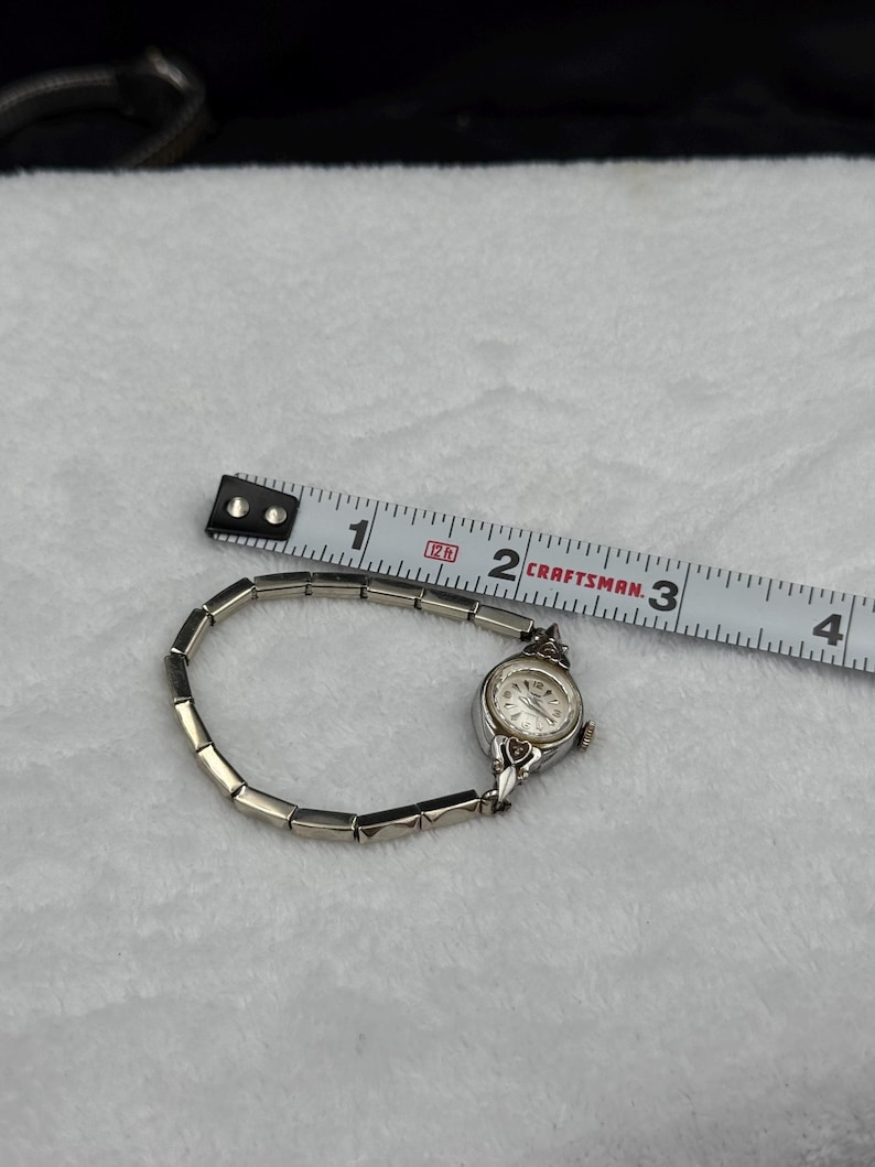 May include: A vintage silver-toned wristwatch with a small, round face and a linked band. The watch is next to a measuring tape, showing a length of approximately 3 inches. The watch face has a white background with black numerals and hands.