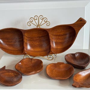 Pode incluir: Um conjunto de travessas de madeira, incluindo uma grande bandeja de três secções e várias tigelas menores de diferentes formas. A madeira tem um tom castanho-avermelhado quente. A bandeja é suportada por um suporte de metal dourado.