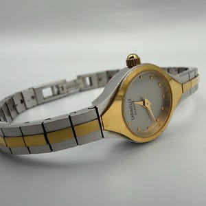 May include: A two-tone wristwatch featuring a silver and gold-colored metal link band. The round watch face is silver with gold-colored hands and hour markers. The text CARAVELLE QUARTZ is printed on the face. A gold-colored crown is on the side.