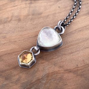 Puede incluir: Un collar de plata con un colgante triangular nacarado y un colgante hexagonal de piedra amarilla. La cadena es de color plata oscura y los colgantes están engastados en plata. El collar se exhibe sobre una superficie de madera.