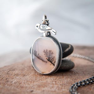 May include: A silver pendant necklace featuring an oval-shaped dendritic agate stone with dark, tree-like inclusions. A small, stylized silver bird figure is attached to the top of the stone setting, which is connected to a delicate silver chain.