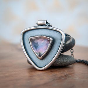 Puede incluir: Un collar con colgante de plata con un diseño triangular. El colgante presenta una piedra preciosa de color púrpura claro engastada en un marco plateado. El collar se exhibe sobre una superficie de madera con un fondo borroso.