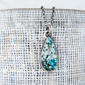 May include: A teardrop-shaped pendant necklace featuring a mottled turquoise stone with shades of light blue, dark blue, and black matrix. The stone is set in a dark metal bezel and hangs from a matching dark metal chain with small circular links.
