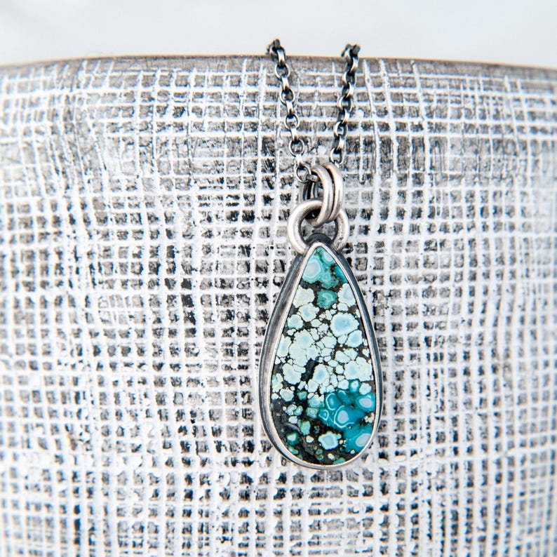 May include: A teardrop-shaped pendant necklace featuring a mottled turquoise stone with shades of light blue, dark blue, and black matrix. The stone is set in a dark metal bezel and hangs from a matching dark metal chain with small circular links.