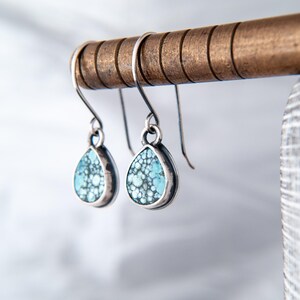May include: A pair of teardrop-shaped earrings featuring light blue and dark grey speckled stone inlays set in polished silver frames. The earrings hang from curved silver ear wires, showcasing a detailed matrix pattern within the stone surface.