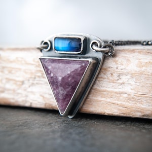 May include: A silver necklace featuring a triangular purple gemstone and a rectangular blue gemstone. The pendant is set in a silver frame and hangs from a delicate chain. The necklace is displayed on a wooden surface.
