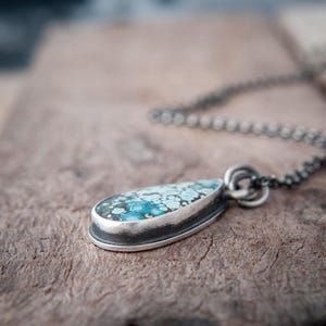 May include: A teardrop-shaped pendant featuring a mottled blue and white stone set in a dark, oxidized silver bezel. The pendant is attached to a matching dark, oxidized silver chain, showcasing a textured, metallic finish throughout the piece.