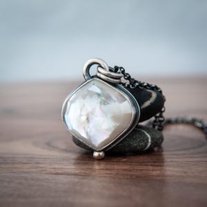 May include: A necklace featuring a faceted, iridescent white mother-of-pearl pendant set in a dark, oxidized silver frame. The pendant is attached to a dark, fine-linked silver chain, creating a contrast between the light stone and metal.