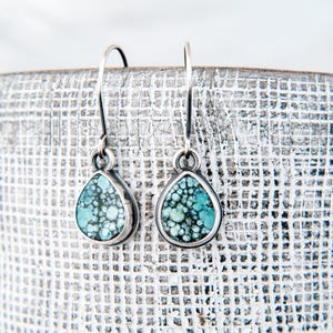 May include: A pair of teardrop-shaped earrings featuring polished turquoise stones with dark matrix patterns set in silver bezels. The earrings hang from simple silver shepherd hook wires, showcasing a natural blue-green color palette.