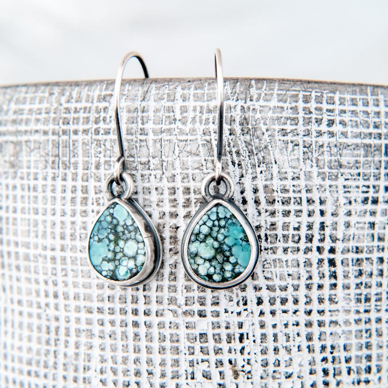 May include: A pair of teardrop-shaped earrings featuring polished turquoise stones with dark matrix patterns set in silver bezels. The earrings hang from simple silver shepherd hook wires, showcasing a natural blue-green color palette.