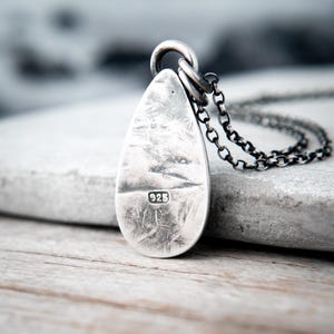 May include: A teardrop-shaped silver pendant with a distressed, textured surface and a dark oxidized finish. The pendant features a stamped 925 hallmark and is attached to a dark, round-link silver chain, creating a cohesive metallic aesthetic.