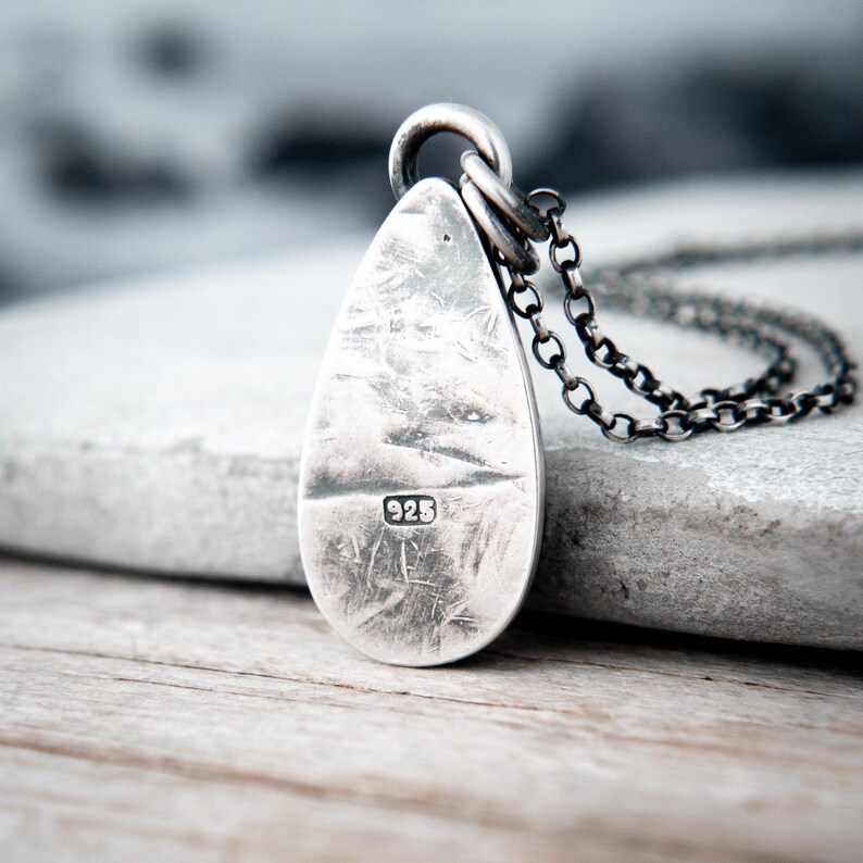 May include: A teardrop-shaped silver pendant with a distressed, textured surface and a dark oxidized finish. The pendant features a stamped 925 hallmark and is attached to a dark, round-link silver chain, creating a cohesive metallic aesthetic.