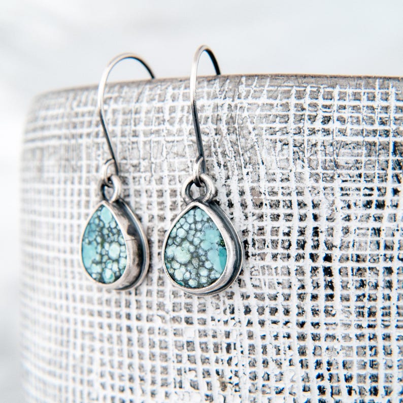 May include: A pair of teardrop-shaped earrings featuring light blue and dark green matrix stone inlays set in oxidized silver. Each earring hangs from a curved silver hook, showcasing a detailed, speckled pattern within the stone and a smooth metal border.