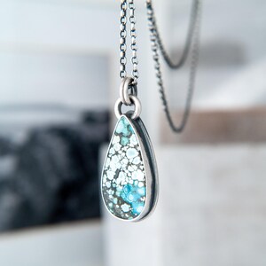 May include: A teardrop-shaped pendant featuring a speckled turquoise stone with blue, white, and dark grey patterns, set in a polished silver bezel. The pendant hangs from a delicate silver chain with round links, showcasing a metallic finish.
