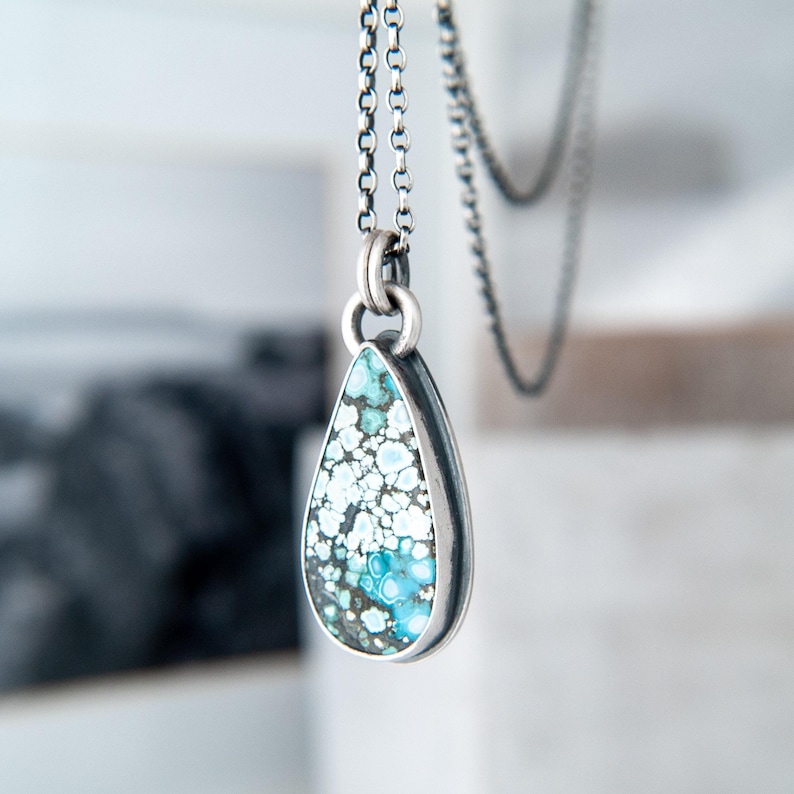 May include: A teardrop-shaped pendant featuring a speckled turquoise stone with blue, white, and dark grey patterns, set in a polished silver bezel. The pendant hangs from a delicate silver chain with round links, showcasing a metallic finish.
