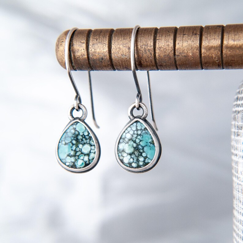 May include: A pair of teardrop-shaped dangle earrings featuring a mottled turquoise and black stone inlay set in a dark silver-toned metal frame. The earrings hang from simple curved ear wires, showcasing a detailed, pebble-like texture.