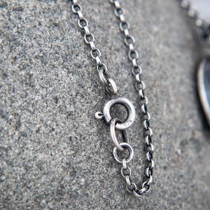 May include: A silver chain necklace featuring a round spring ring clasp. The metal surface has a dark, oxidized finish. The clasp is clearly stamped with the number 925, indicating the sterling silver composition of the jewelry piece.