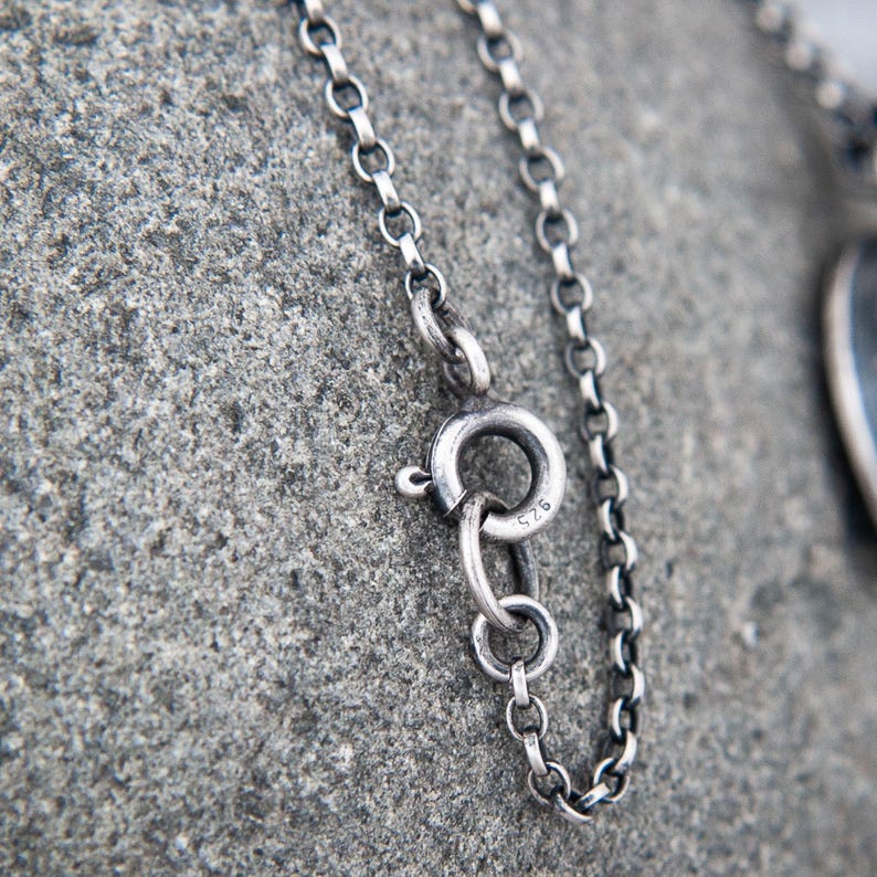 May include: A silver chain necklace featuring a round spring ring clasp. The metal surface has a dark, oxidized finish. The clasp is clearly stamped with the number 925, indicating the sterling silver composition of the jewelry piece.