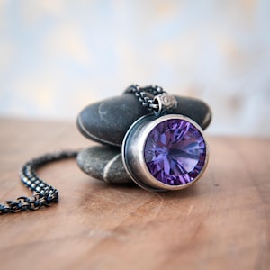 May include: A silver pendant necklace featuring a large, round, faceted purple gemstone. The gemstone is set in a bezel setting and hangs from a dark chain. The necklace rests on a stack of smooth, gray stones.