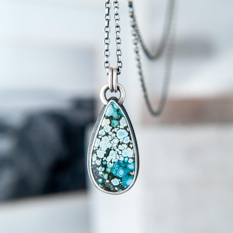 May include: A teardrop-shaped pendant necklace featuring a polished stone with a mottled pattern of light blue, teal, and dark brown matrix. The stone is set in a smooth silver bezel and hangs from a delicate silver chain with round links.