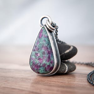 May include: A teardrop-shaped pendant featuring a mottled green and magenta stone set in a dark, oxidised silver bezel. The pendant is attached to a matching dark silver chain, with the metal showing a textured, rustic finish throughout the piece.