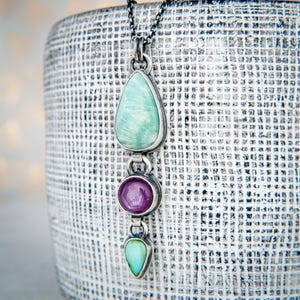 May include: A silver pendant necklace featuring three gemstone pendants. The top pendant is a light green teardrop shape, the middle is a round purple stone, and the bottom is a small green teardrop. The necklace hangs on a silver chain.