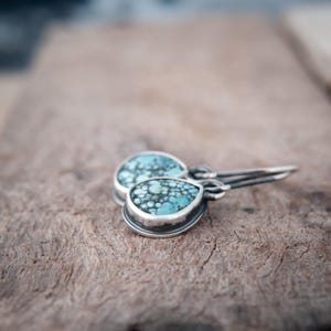 May include: A pair of teardrop-shaped earrings featuring speckled turquoise stones set in oxidized silver bezels. The earrings have silver ear wires and display a distinct matrix pattern of blue, green, and dark grey tones within the stone surface.