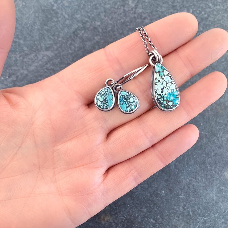 May include: A jewelry set consisting of a teardrop-shaped pendant on a silver chain and matching teardrop-shaped dangle earrings. Each piece features a speckled turquoise stone with dark matrix patterns set in a polished silver frame.