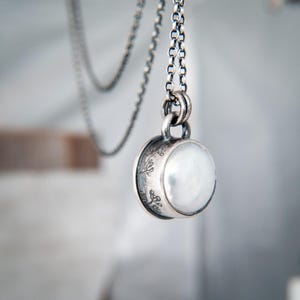 May include: A round pendant necklace featuring a smooth, white, iridescent stone set in a dark, oxidized silver casing. The side of the silver setting is decorated with a delicate, etched floral pattern. The pendant hangs from a matching silver chain.