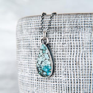 May include: A teardrop-shaped pendant featuring a mottled turquoise stone with intricate blue and black matrix patterns, set in a dark oxidized silver bezel. The pendant hangs from a matching dark silver chain with small, rounded links.