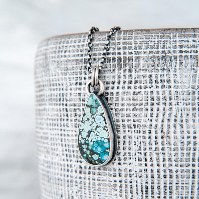 May include: A teardrop-shaped pendant featuring a mottled turquoise stone with intricate blue and black matrix patterns, set in a dark oxidized silver bezel. The pendant hangs from a matching dark silver chain with small, rounded links.