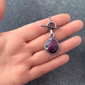 May include: A pendant necklace featuring a faceted, teardrop-shaped magenta and dark grey stone set in a silver-toned metal frame. The pendant is attached to a silver-toned toggle clasp mechanism consisting of a circular ring and a horizontal bar.