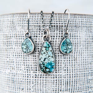 May include: A jewelry set consisting of a necklace and two matching drop earrings. Each piece features a teardrop-shaped stone with a mottled pattern of light blue, teal, and dark grey, set in a dark oxidized silver frame with a chain.