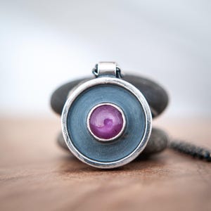 May include: A circular pendant featuring a smooth, cabochon-cut magenta gemstone set in the centre of a dark, oxidised silver disc. The pendant has a polished silver rim and a small, rectangular bail at the top for attaching to a chain.