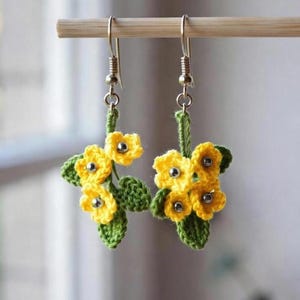 Può includere: Orecchini all'uncinetto fatti a mano con grappoli di fiori gialli, centri argentati e foglie verdi. Gli orecchini sono sospesi a ganci color argento, mostrando un delicato design floreale.