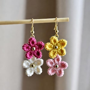 以下が含まれることがあります： かぎ針編みの花のイヤリング。片方のイヤリングはマゼンタと白の花、もう片方は黄色とピンクの花です。各花は金色の中心を持ち、金色のフックから吊り下げられています。