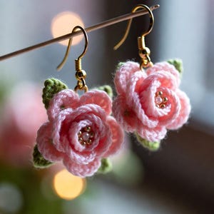 Könnte beinhalten: Ein Paar handgefertigte Häkelohrringe in Form von rosa Rosen mit goldfarbenen Haken. Die Rosenblätter sind zartrosa, mit grünen Blättern am Ansatz. Die Mitte jeder Blume hat kleine, goldfarbene Perlen.