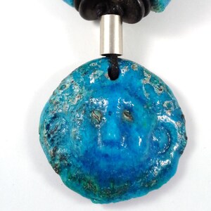May include: A turquoise pendant necklace with a smiling face design. The pendant is a vibrant blue with a textured surface and silver accents. The necklace has a black cord and a silver metal tube.