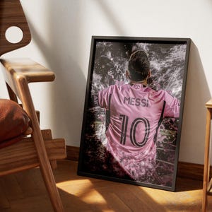 May include: A black framed poster with a pink and black image of a soccer player wearing a pink jersey with the number 10 and the name "Messi" on the back. The player is surrounded by a white and gray abstract design.
