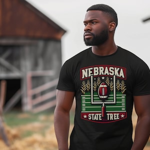May include: Black t-shirt with a Nebraska football design. The graphic includes a football, goalpost, and the words "NEBRASKA STATE TREE" in a rectangular frame. The design is in red, white, and green.