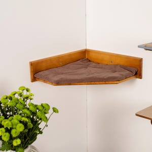 Puede incluir: Estante esquinero de madera para gatos con cojín marrón. El estante está montado en una pared blanca. El estante es triangular y tiene un borde elevado. El cojín es de color marrón liso. Un jarrón con flores verdes está en primer plano.