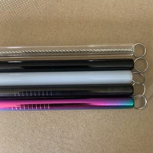 May include: A collection of reusable drinking straws in various colors and materials. Includes a clear glass straw with a cleaning brush, a black straw, a white straw, a dark grey straw, and a rainbow-colored straw, each with a small metal ring.