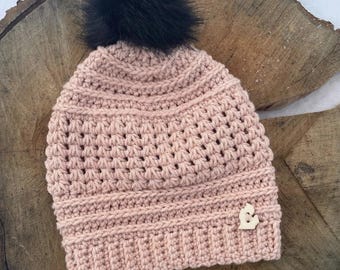 May include: A pink crocheted beanie with a black faux fur pom pom and a wooden charm. The beanie has a textured stitch pattern.