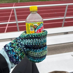 Pode incluir: Uma garrafa de pl&aacute;stico transparente de Mountain Dew com uma tampa amarela, segurada em uma luva de croch&ecirc; azul e verde.