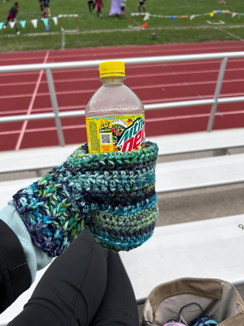 Pode incluir: Uma garrafa de pl&aacute;stico transparente de Mountain Dew com uma tampa amarela, segurada em uma luva de croch&ecirc; azul e verde.