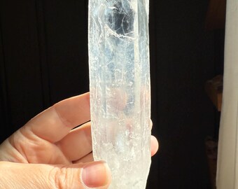 May include: A clear quartz crystal point, held in a hand, with a faceted top and a rough, natural base. The crystal is translucent, allowing light to pass through, and has internal inclusions. The background is dark, with sunlight illuminating the crystal.